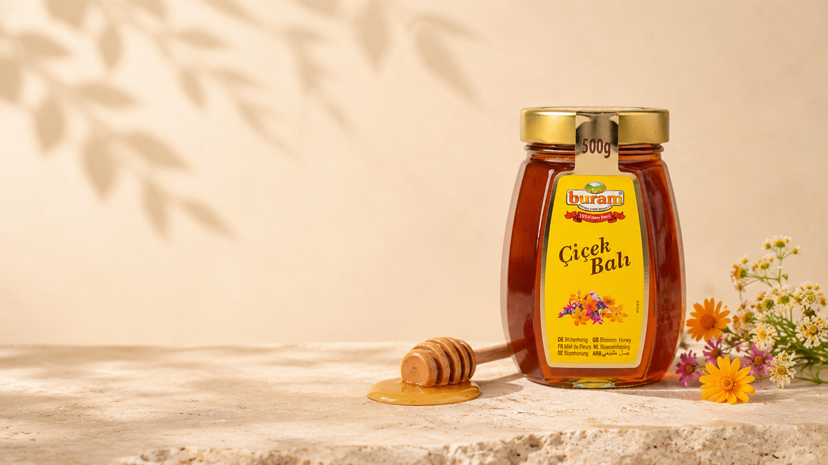 Buram Blütenhonig im Glas, daneben ein Honiglöffel und Wildblumen auf hellem Stein